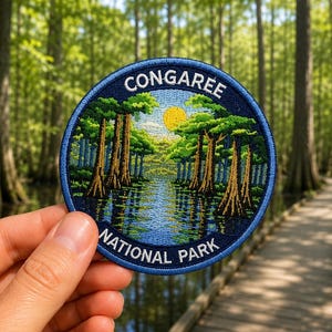 May include: A circular embroidered patch featuring a landscape of Congaree National Park. The design depicts a swampy waterway lined with tall cypress trees, a bright yellow sun, and blue sky. The text CONGAREE NATIONAL PARK is stitched in white.