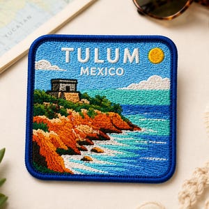 May include: A square embroidered patch featuring a coastal landscape of Tulum, Mexico. The design shows a stone ruin on a cliffside overlooking a turquoise ocean with white waves, a yellow sun, and white clouds. The text TULUM MEXICO is stitched in white.