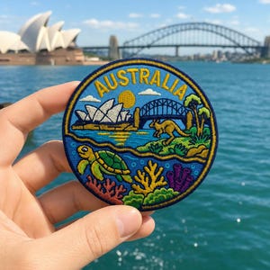 May include: A circular embroidered patch featuring the word AUSTRALIA in yellow text. The design depicts a blue sky with a yellow sun, the Sydney Opera House, the Sydney Harbour Bridge, a kangaroo, and an underwater scene with a sea turtle.