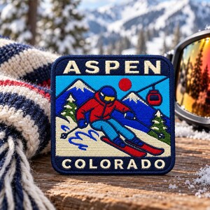 May include: A square embroidered patch featuring a skier in a red jacket and blue pants on a snowy mountain. The text ASPEN is at the top and COLORADO is at the bottom in white lettering. A red sun and a ski lift gondola are visible in the blue sky.