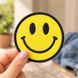 May include: A circular embroidered patch featuring a bright yellow smiley face with black stitched eyes and a wide black curved mouth. The patch has a thick black border around the entire circumference, creating a bold and high-contrast design.
