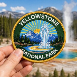 May include: A circular embroidered patch featuring a landscape of Yellowstone National Park. The design includes a geyser erupting, blue mountains, green pine trees, and a yellow sun with rays, all bordered by a dark green ring with yellow text.
