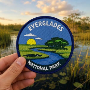 May include: A circular embroidered patch featuring a landscape of the Everglades National Park. The design includes a blue sky with a yellow sun, green marshland, a winding blue river, and a black alligator, with the text EVERGLADES NATIONAL PARK.