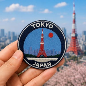 May include: A circular embroidered patch featuring a light blue sky, a red Tokyo Tower, a red sun, and a faint city skyline. The dark blue border contains the white text TOKYO at the top and JAPAN at the bottom, with a thin white inner ring.