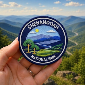 May include: A circular embroidered patch featuring a landscape of Shenandoah National Park. The design includes blue mountains, a winding river, green trees, a yellow sun, and white clouds. The text SHENANDOAH NATIONAL PARK is stitched in white.