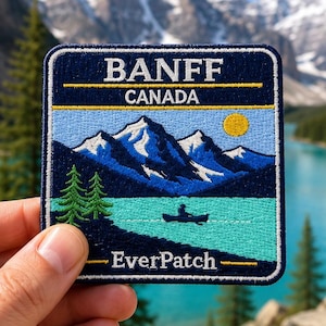 May include: A square embroidered patch featuring a landscape of Banff, Canada. The design includes dark blue mountains with white snow caps, a turquoise lake, a yellow sun, and a person in a canoe. The text reads BANFF, CANADA, and EverPatch.
