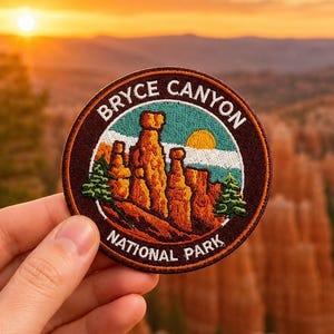 Może przedstawiać: Okrągła haftowana naszywka przedstawiająca krajobraz Parku Narodowego Bryce Canyon z pomarańczowymi formacjami skalnymi, zielonymi sosnami i żółtym słońcem na tle turkusowego nieba, otoczona ciemnobrązową obwódką z napisem Bryce Canyon National Park.