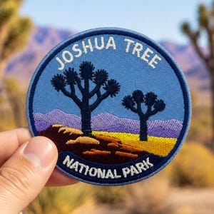 Puede incluir: Un parche bordado circular que muestra un paisaje desértico con dos siluetas oscuras de árboles de Josué sobre un cielo azul claro. El texto JOSHUA TREE NATIONAL PARK está bordado en blanco a lo largo de los bordes superior e inferior del borde azul.