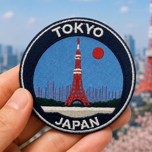 May include: A circular embroidered patch featuring a light blue sky, a red Tokyo Tower, a red sun, and a city skyline. The dark blue border contains the white text TOKYO at the top and JAPAN at the bottom, with a thin white inner circular trim.