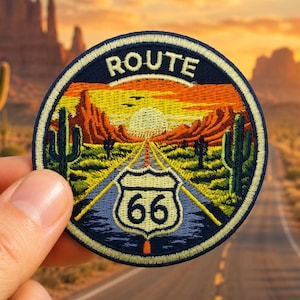 May include: A circular embroidered patch featuring a desert landscape with a road leading toward a sunset, saguaro cacti, and a Route 66 shield sign. The text ROUTE is stitched in white at the top, and the number 66 is centered on the shield.