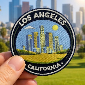May include: A circular embroidered patch featuring a stylized Los Angeles city skyline in shades of yellow and blue against a light blue sky with a yellow sun. The text LOS ANGELES and CALIFORNIA is stitched in white along the dark blue border.