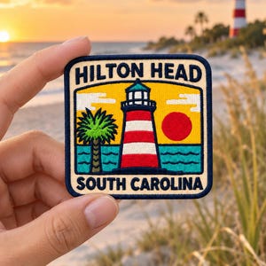 Peut inclure: Un écusson brodé carré représentant un phare rayé rouge et blanc, un palmier vert et un soleil rouge au-dessus d'une mer bleue. Le texte HILTON HEAD figure en haut et SOUTH CAROLINA en bas, le tout bordé par un cadre bleu foncé.