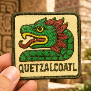 May include: A square embroidered patch with a cream-colored background and a dark green border. The design features a green serpent with red crest feathers, a yellow eye, and a red tongue. The text QUETZALCOATL is embroidered in dark green letters.