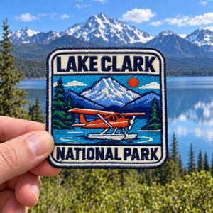 May include: A square embroidered patch featuring a bright orange floatplane on blue water, with a snow-capped mountain, green pine trees, and a red sun in the background. The text LAKE CLARK is at the top and NATIONAL PARK is at the bottom in dark blue.