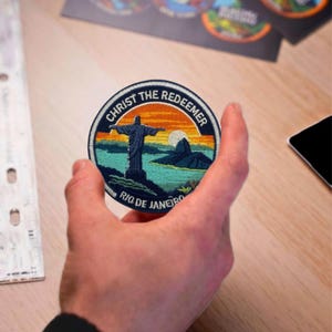以下が含まれることがあります： リオデジャネイロのキリスト像を描いた、円形の刺繍パッチ。「CHRIST THE REDEEMER」と「RIO DE JANEIRO」の文字入り。像、山、水上の夕焼けが描かれており、青、オレンジ、緑の色合いです。