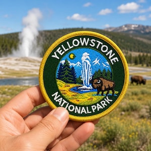 Op de afbeelding: Een ronde geborduurde patch met de tekst "YELLOWSTONE NATIONAL PARK" in geel en wit. De patch heeft een groene rand en een blauw midden met een geiser, bizon, bomen en bergen.