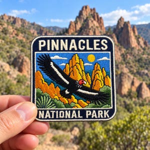 May include: A rectangular embroidered patch featuring a black condor with a red head flying over yellow rock formations and green vegetation. The sky is blue with a yellow sun and white clouds. The text PINNACLES NATIONAL PARK is in white.