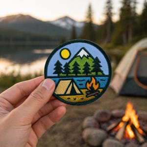 May include: A round embroidered patch featuring a camping scene with a tent, campfire, trees, lake, mountain, and sun. The patch has a blue border and is held in a hand. The background shows a lake, mountains, and a tent.