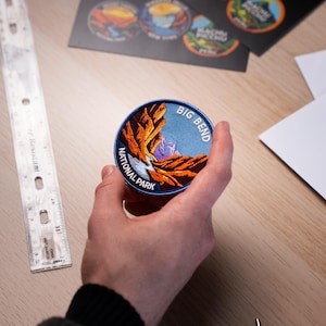 Puede incluir: Un parche bordado redondo con una escena paisajística del Parque Nacional Big Bend. El parche tiene un borde azul con las palabras "BIG BEND NATIONAL PARK". La imagen representa un cañón con tonos naranjas y morados sobre un cielo azul.