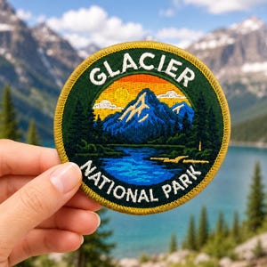 Op de afbeelding: Ronde geborduurde patch met de tekst "GLACIER NATIONAL PARK". De patch toont een berglandschap met een meer, bomen en een zonsondergang. Het heeft een gouden rand en wordt in een hand gehouden.