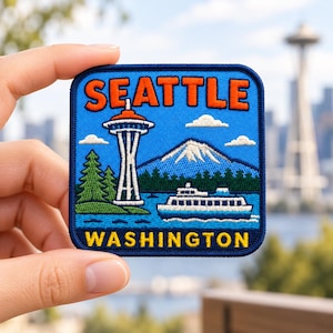 Puede incluir: Un parche bordado cuadrado con fondo azul, el texto naranja SEATTLE en la parte superior y el texto amarillo WASHINGTON en la parte inferior. El diseño muestra la Space Needle, una montaña nevada, pinos verdes y un ferry blanco sobre el agua.