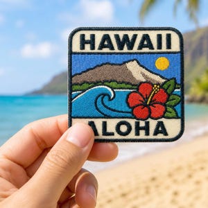 Op de afbeelding: Een vierkante geborduurde patch met de tekst HAWAII bovenaan en ALOHA onderaan. Het ontwerp toont een bruine berg, een gele zon, een blauwe oceaangolf en een grote rode hibiscusbloem met groene bladeren op een crèmekleurige achtergrond.
