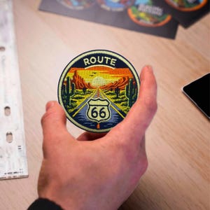 May include: A circular embroidered patch featuring a Route 66 design. The patch depicts a desert landscape with a road, cacti, and a setting sun. The word "ROUTE" is arched across the top, and "66" is in a shield shape.