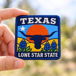 May include: A square embroidered patch featuring a dark blue border and the text TEXAS at the top and LONE STAR STATE at the bottom. The design shows a dark blue longhorn silhouette against an orange sun, a white star, and blue flowers.