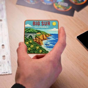 May include: Embroidered patch featuring a scenic view of Big Sur, California. The design includes a bridge, coastline, ocean, and the text "BIG SUR CALIFORNIA". The patch has a blue border and a yellow sun.