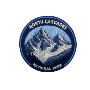 May include: Embroidered patch featuring a mountain range scene. The patch is circular with a dark blue border and a light blue background. The text "NORTH CASCADES NATIONAL PARK" is embroidered around the edge in white.