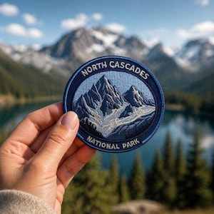 Può includere: Una toppa rotonda ricamata con un disegno di catena montuosa e la scritta "NORTH CASCADES NATIONAL PARK". La toppa ha un bordo blu e uno sfondo azzurro con dettagli di montagne bianche e blu navy.
