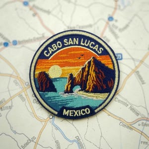 May include: Circular embroidered patch depicting a Cabo San Lucas, Mexico, landscape with a sunset over the sea and rock formations. The patch has a navy blue border with the text "CABO SAN LUCAS" and "MEXICO".