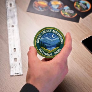 Puede incluir: Un parche redondo bordado con el logotipo del Parque Nacional Great Smoky Mountains. El parche tiene un borde verde con texto, una escena de montaña azul y verde, una luna creciente y estrellas.