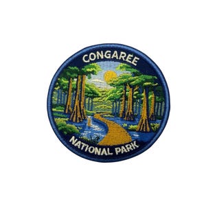 May include: Embroidered patch featuring a scenic view of Congaree National Park. The design includes a yellow path winding through a forest with tall trees, a river, and a sun. The patch is round with a navy blue border and the text "CONGAREE NATIONAL PARK".