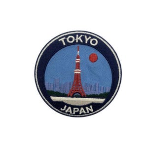 May include: Embroidered patch featuring the Tokyo Tower in red and white, set against a blue sky with a red sun. The patch is circular, with the words "TOKYO" and "JAPAN" in white lettering on a navy blue border.