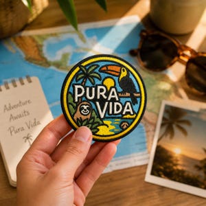 May include: Round embroidered patch with the words "Pura Vida" and a toucan, sloth, and palm trees. The patch has a blue, yellow, and black border. A notebook with the words "Adventure Awaits Pura Vida" is in the background.