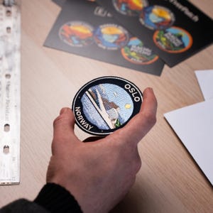 May include: A round embroidered patch featuring a scenic view of Oslo, Norway, with the city's name in white lettering. The patch has a black border and depicts a mountain, water, and sky. Other patches and cards are in the background.