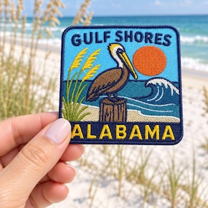 Peut inclure: Un écusson brodé carré représentant un pélican brun perché sur un poteau en bois. L'image montre des avoines de mer jaunes, un océan bleu avec une vague blanche et un soleil orange. Le texte GULF SHORES figure en haut et ALABAMA en bas.