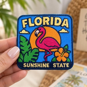 May include: A square embroidered patch featuring a bright pink flamingo standing in front of a large orange sun. The design includes green palm leaves, an orange hibiscus flower, and the text FLORIDA and SUNSHINE STATE in yellow letters.