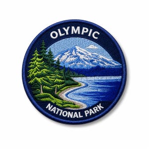Op de afbeelding: Geborduurde ronde patch met een landschap van Olympic National Park. Het ontwerp toont een besneeuwde berg, een meer en groenblijvende bomen, alles binnen een marineblauwe rand. De woorden "OLYMPIC NATIONAL PARK" zijn op de patch geborduurd.