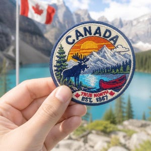 Könnte beinhalten: Ein runder gestickter Aufnäher mit einer Landschaftsszene, die einen dunkelblauen Elch, ein rotes Kanu, eine gelbe Sonne und weiße Berge zeigt. Der Text lautet oben CANADA und unten TRUE NORTH EST. 1867, ergänzt durch kleine rote Ahornblatt-Symbole.