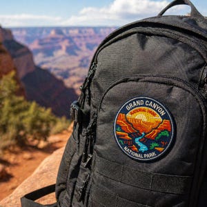 Può includere: Zaino nero con una toppa del Grand Canyon National Park. La toppa presenta un'illustrazione colorata del paesaggio del canyon con le parole "Grand Canyon National Park". Lo zaino ha più scomparti con cerniera.