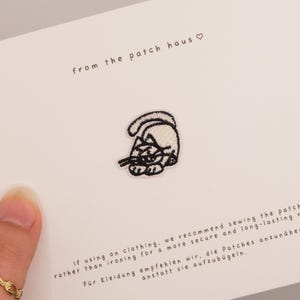Könnte beinhalten: Gestickter Aufnäher mit einer stilisierten Katze in Schwarz und Weiß, mit dem Text "from the patch haus" darüber. Der Aufnäher befindet sich auf einer weißen Karte mit Anweisungen auf Englisch und Deutsch.