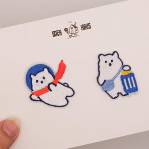 Könnte beinhalten: Zwei gestickte Aufnäher mit Cartoon-Eisbären. Einer trägt einen blauen Weltraumhelm und einen roten Schal, der andere trägt eine blaue Tasche. Die Aufnäher befinden sich auf einer weißen Karte mit dem Text "The Patch Haus".