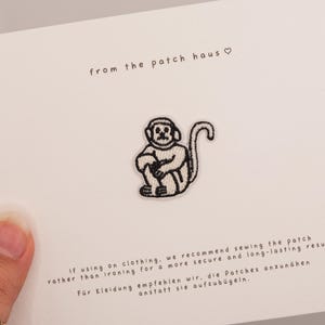 Può includere: Una toppa a forma di scimmia ricamata in bianco e nero su una carta bianca. La carta riporta la scritta "from the patch haus" e le istruzioni in inglese e tedesco. La scimmia è seduta con le braccia attorno alle ginocchia.