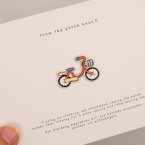 May include: An orange bicycle patch with black wheels, a blue seat, and a basket. The text "from the patch haus" is above the patch. The text below recommends sewing the patch for a secure and long-lasting result.