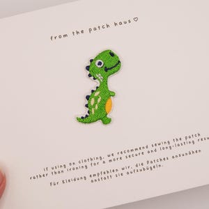 Könnte beinhalten: Ein grüner Dinosaurier-Aufnäher mit einem lächelnden Gesicht, schwarzen Akzenten und einem orangefarbenen Bauch ist auf einer weißen Karte zentriert. Der Text oben lautet "from the patch haus". Darunter wird empfohlen, den Aufnäher für Kleidung aufzunähen.