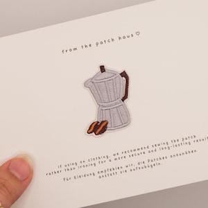 Könnte beinhalten: Eine weiße Karte mit dem Text "from the patch haus" und einem kleinen gestickten Aufnäher einer silbernen Kaffeemaschine mit braunen Akzenten und zwei Kaffeebohnen. Die Karte enthält auch Anweisungen.