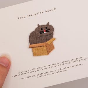 Op de afbeelding: Geborduurde patch met een grijze kat in een bruine doos. De kat heeft een rode neus en zwarte snorharen. De tekst "from the patch haus" staat boven de kat. Instructies staan eronder gedrukt.