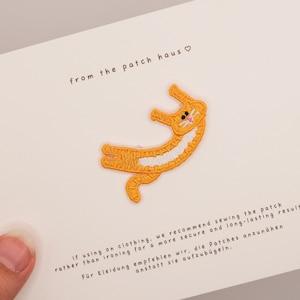 Può includere: Una toppa a forma di gatto ricamato arancione e bianco su una carta bianca. La toppa è in posa giocosa con pancia bianca e naso rosa. Il testo sulla carta recita "from the patch haus" e istruzioni per l'applicazione.